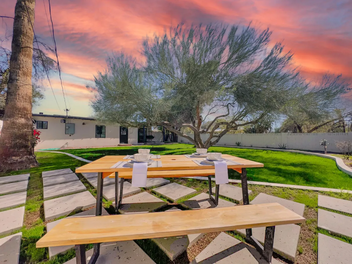 This image captures a spacious backyard with a wooden dining table set on a lush green lawn. A large tree provides natural shade, and the colorful sky suggests the beauty of Arizona sunsets. The scene feels inviting, perfect for outdoor dining, relaxation, and gatherings.