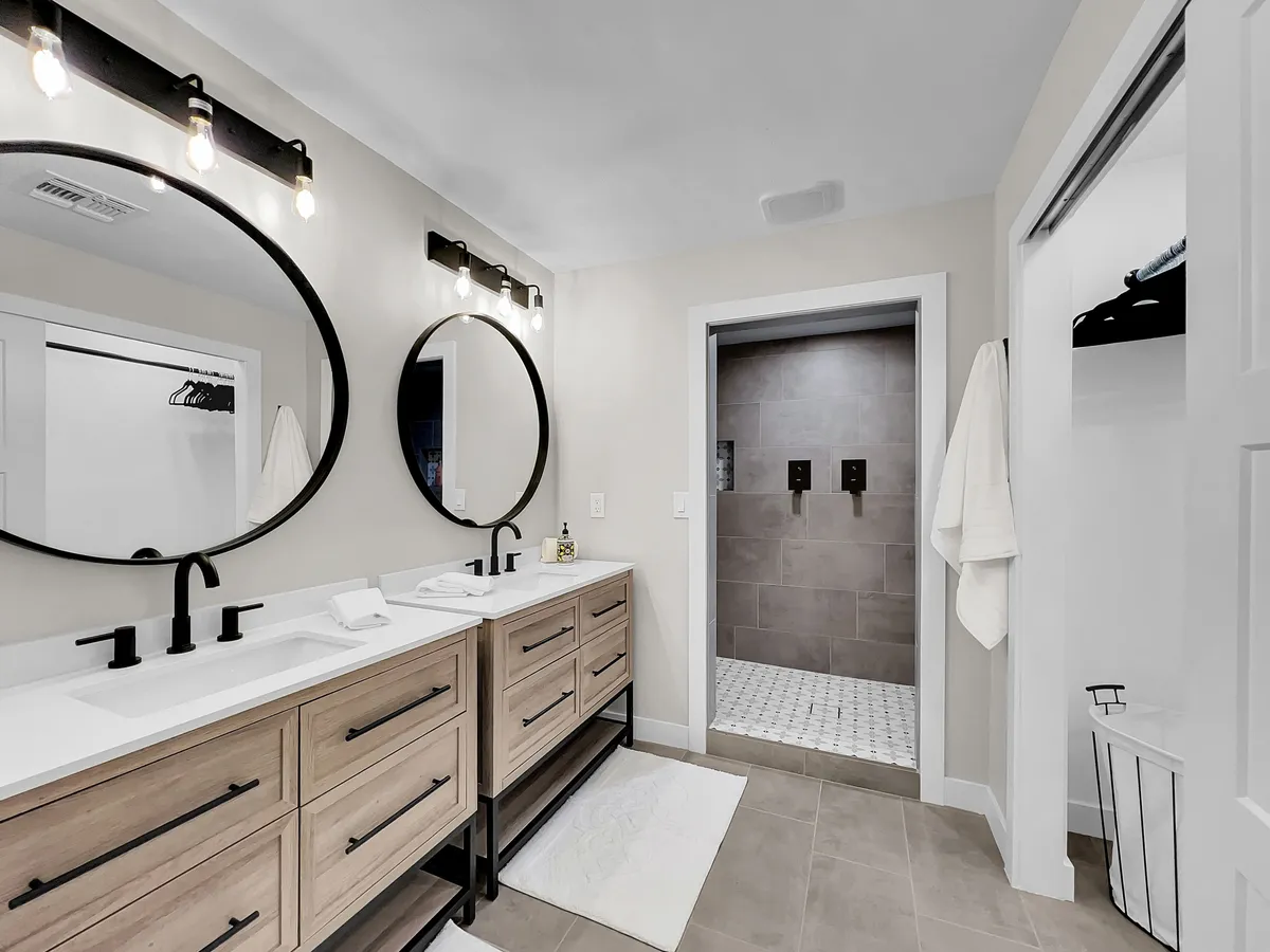 A bright, fresh bathroom featuring a double vanity with mirrors, a spacious shower area, and elegant light fixtures. The room is styled for both practicality and relaxation, evoking a spa-inspired feel.
