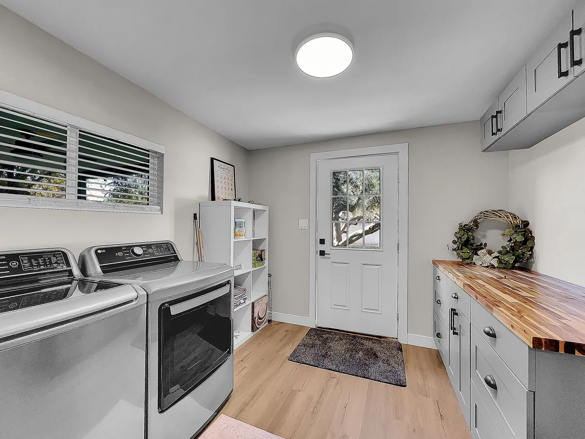 A bright, functional laundry area featuring a washing machine and drying machine beneath a wooden countertop. An open door and neatly arranged shelf unit make the space feel welcoming and useful, supporting day-to-day routines while traveling.
