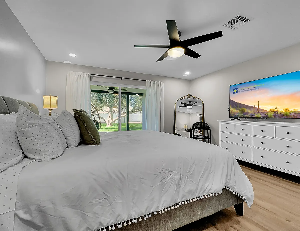 A serene bedroom featuring a large bed with decorative pillows, a sliding glass door that opens to an outdoor view, a TV mounted on the wall, a stylish dresser, and a mirror with a decorative frame. Soft morning sunlight filters in, creating a tranquil atmosphere ideal for relaxation or enjoying a quiet moment.