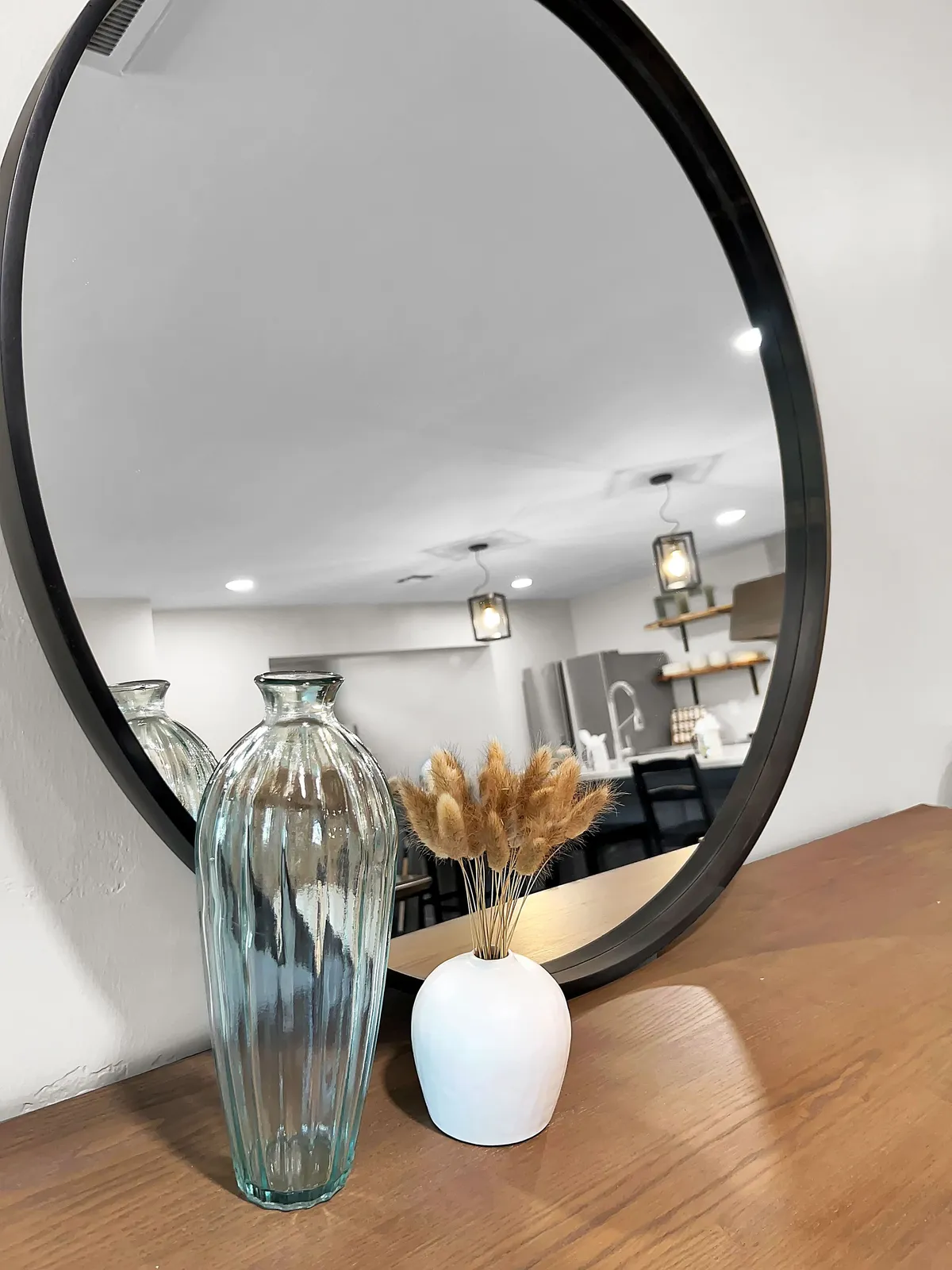 An inviting interior space showcasing a round mirror above a table adorned with a glass and white vase featuring dried flowers. The setting exudes warmth and sophistication, suggesting a well-maintained and thoughtfully designed entryway.